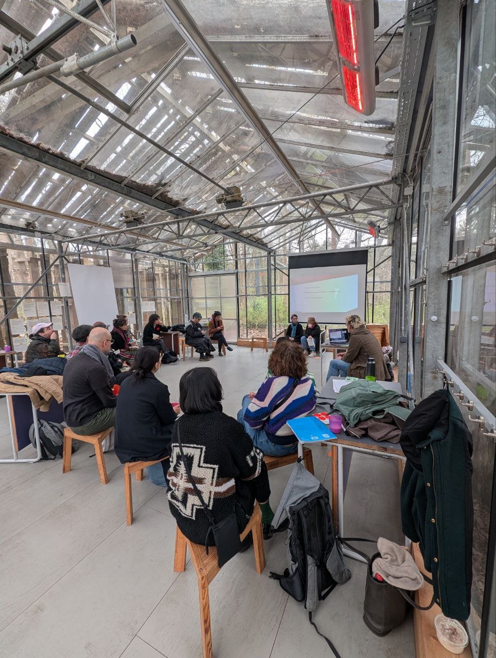 Eine Gruppe von Personen sitzt auf Stühlen im Kreis in einem Haus aus Glas. Auf einer Leinwand ist eine Präsentation zu sehen.